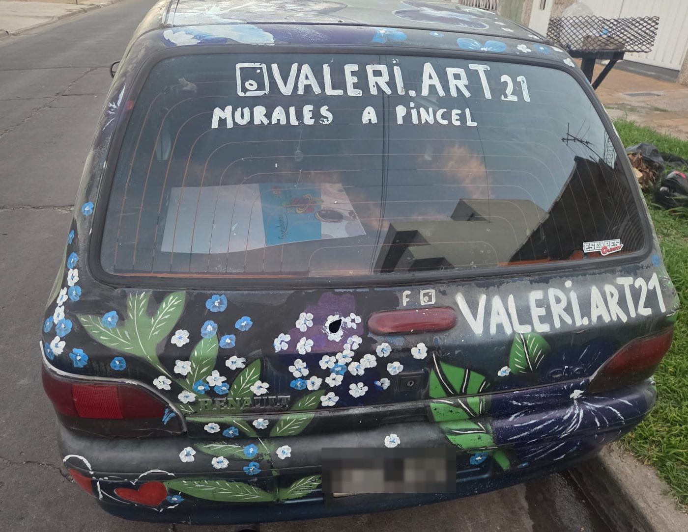 La parte de atrás del mismo auto, con florcitas blancas y celestes y, sobre la luneta, la leyenda «valeri.art21 · murales a pincel».