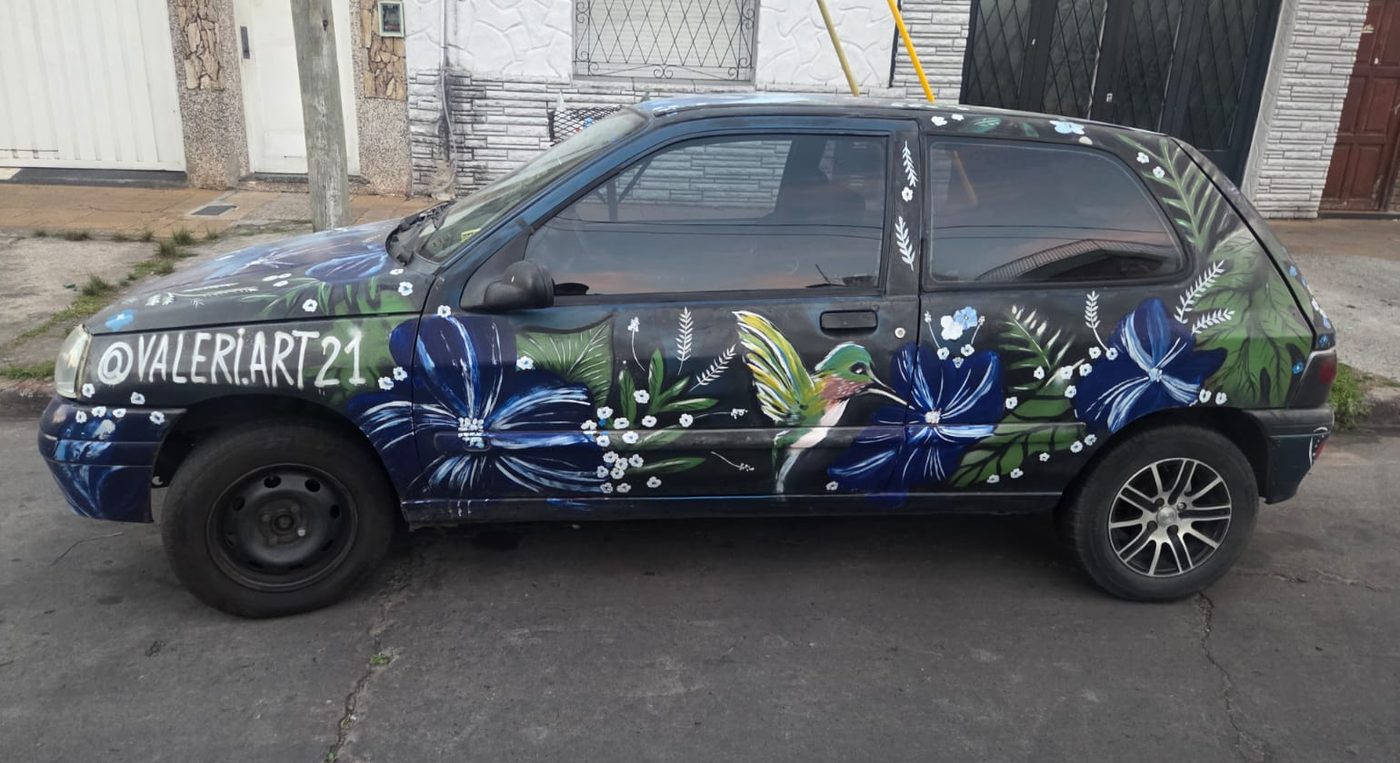 Renault Clio negro pintado a mano de lado a lado con flores azules, helechos y un colibrí, y el nombre @valeri.art21 sobre el capot.