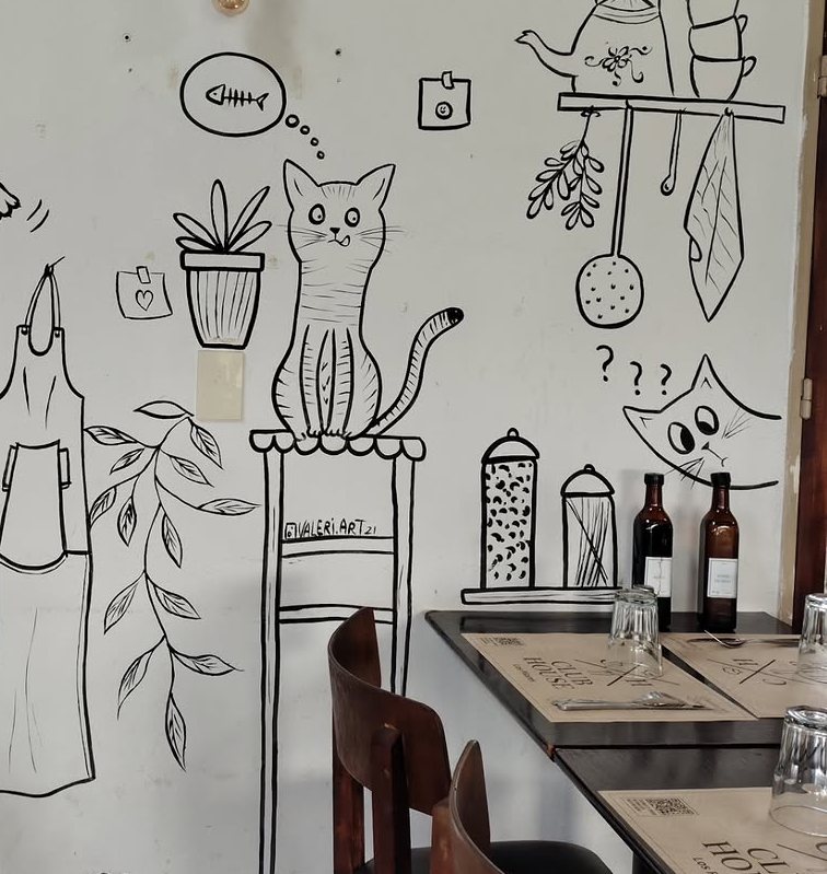 Salón de un bar con las paredes dibujadas en línea negra: gatos, plantas colgantes y objetos de cocina.