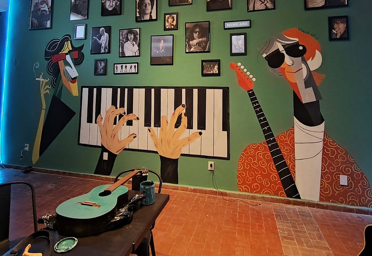 Pared verde de Espacio Montana con un piano pintado en grande, dos manos sobre las teclas y dos músicos caricaturizados, entre cuadros de fotos.