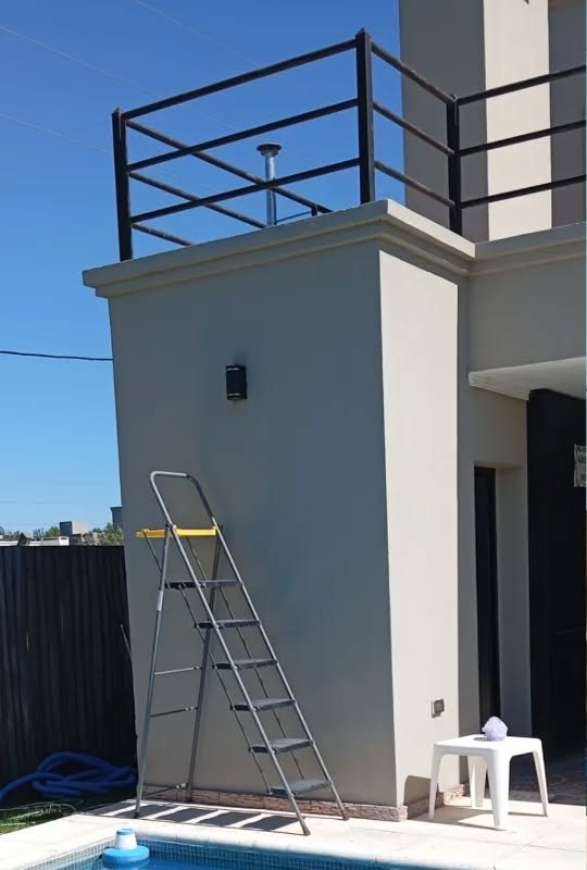 Pared del tanque de agua junto a la pileta, gris y sin pintar, con una escalera apoyada.