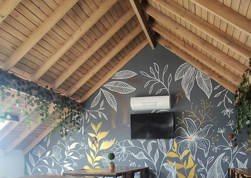 Salón con techo de madera a dos aguas y las paredes pintadas de hojas grandes en gris y amarillo.