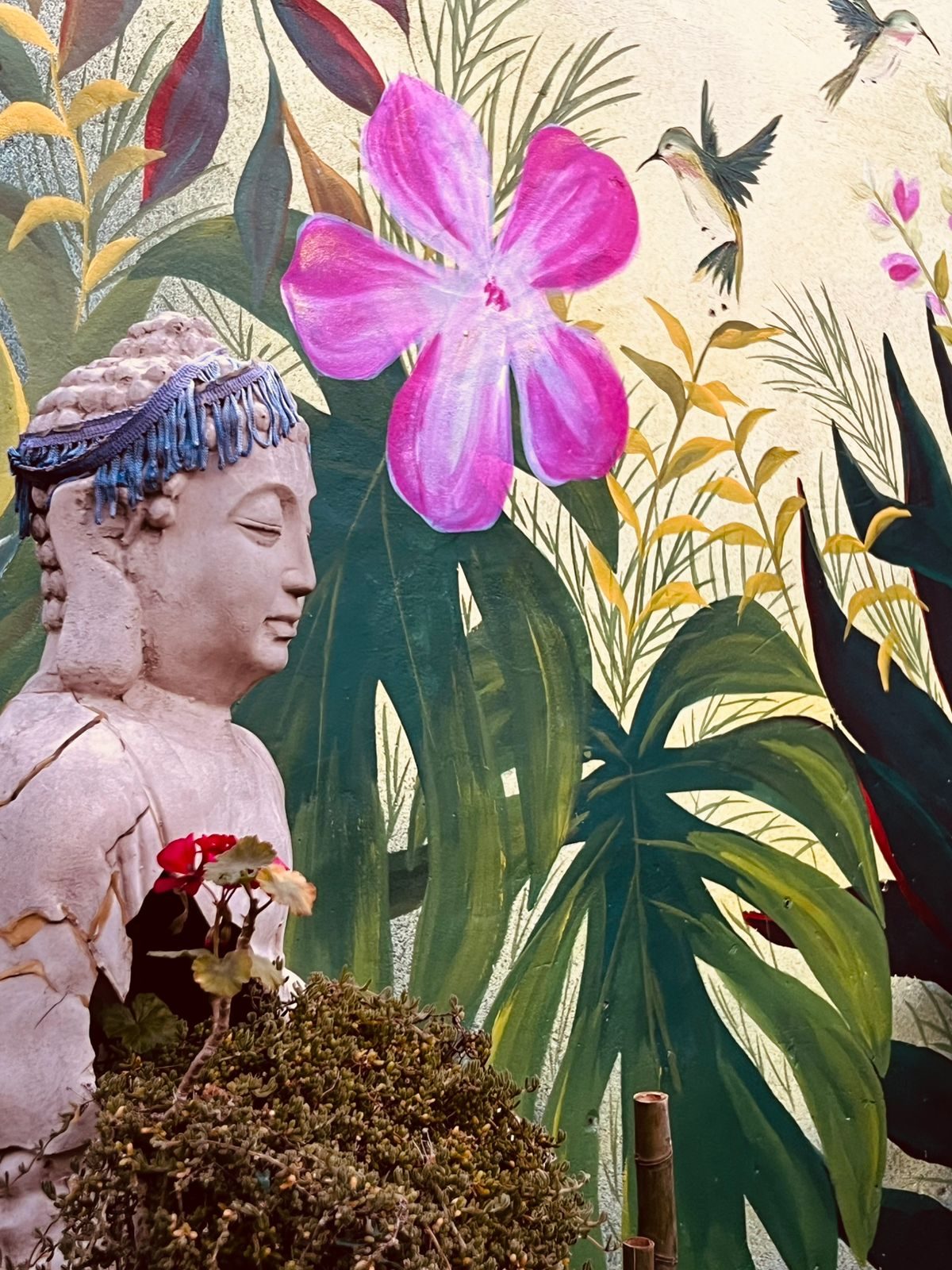 Mural de hibisco rosa, hojas tropicales y colibríes sobre pared clara, con una escultura delante.