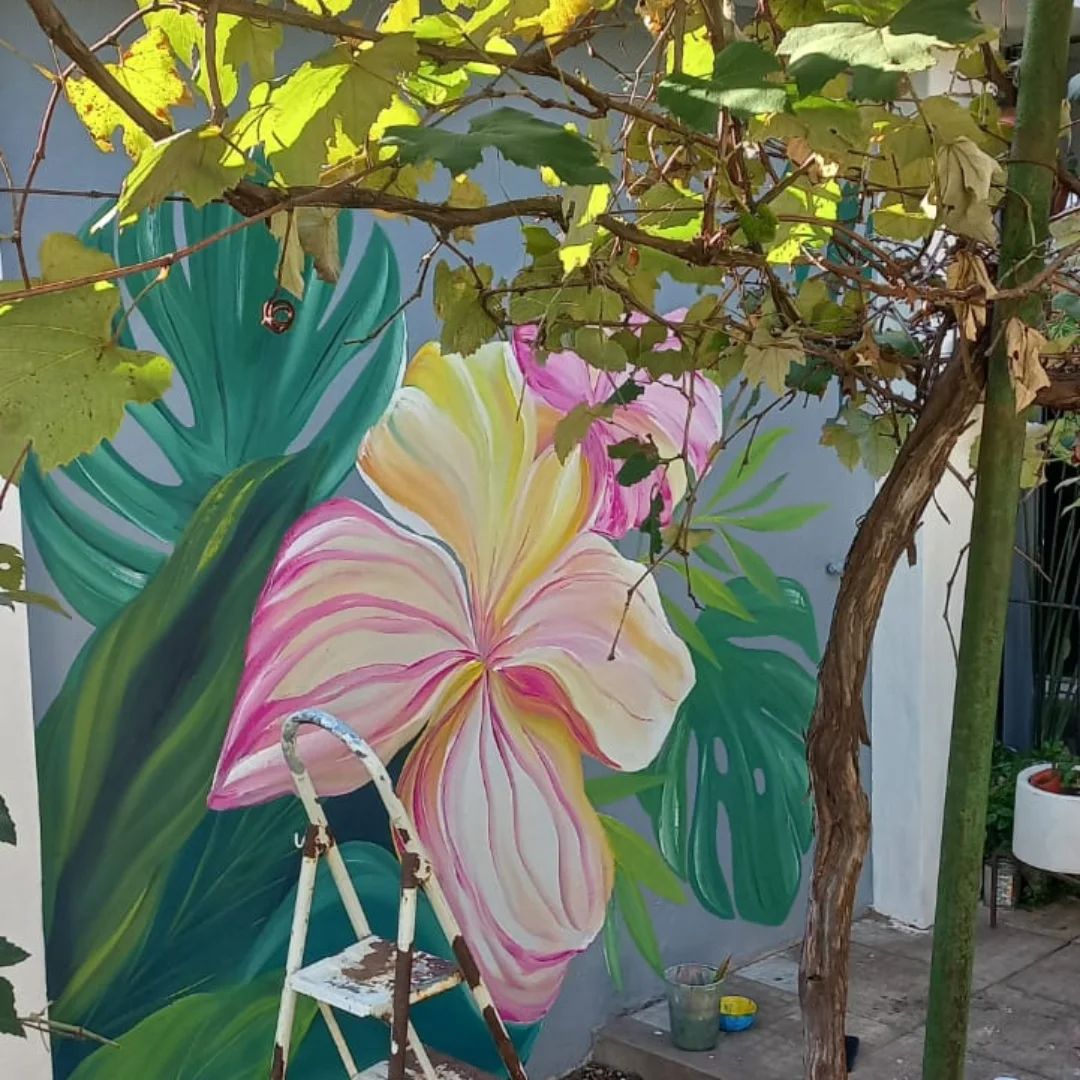 Muro de patio con un hibisco rosa y amarillo entre hojas de monstera, bajo una parra.