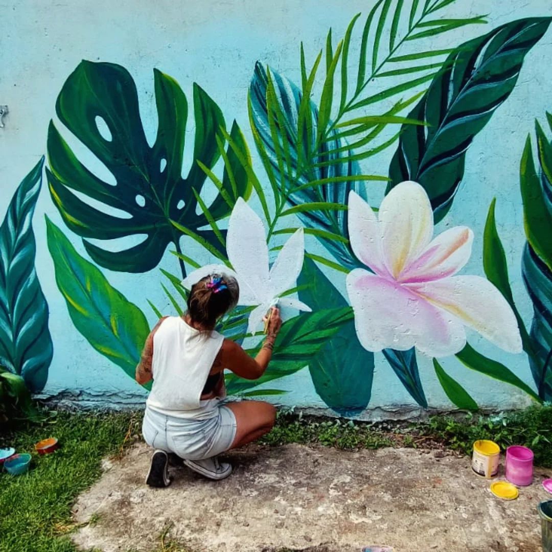 Valeria en cuclillas pintando una flor blanca entre monsteras sobre un muro turquesa, junto a la pileta.