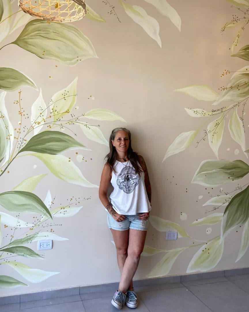 Valeria sonriendo, parada frente al mural de hojas verdes con detalles dorados que pintó para Café Loreto.